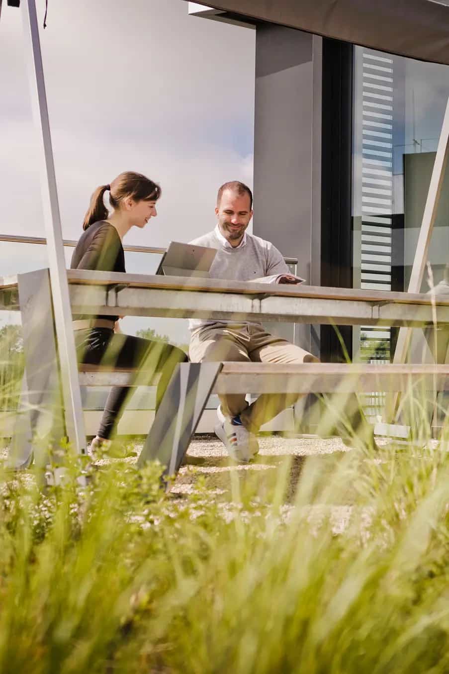 Weibliche und männliche Person sitzen draußen am Tisch auf einer begrünten Dachterrasse.