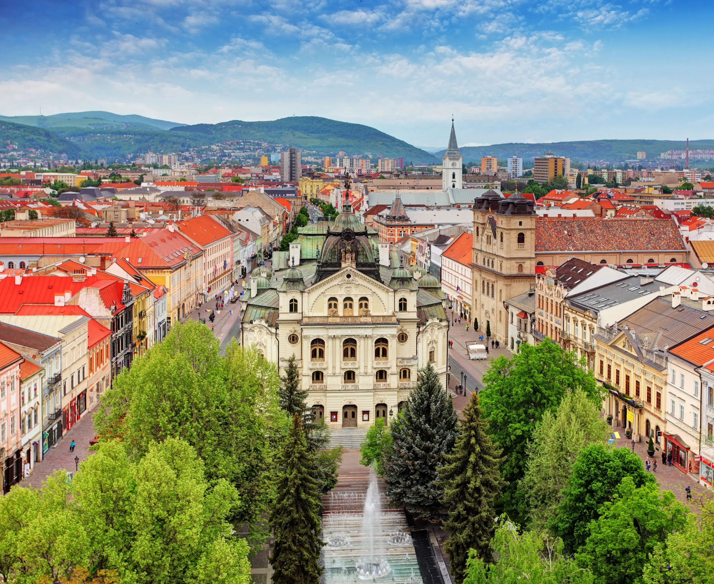 Historisches Stadtzentrum von Kosice mit Gebäuden, Kirche und umliegenden Hügeln unter blauem Himmel.